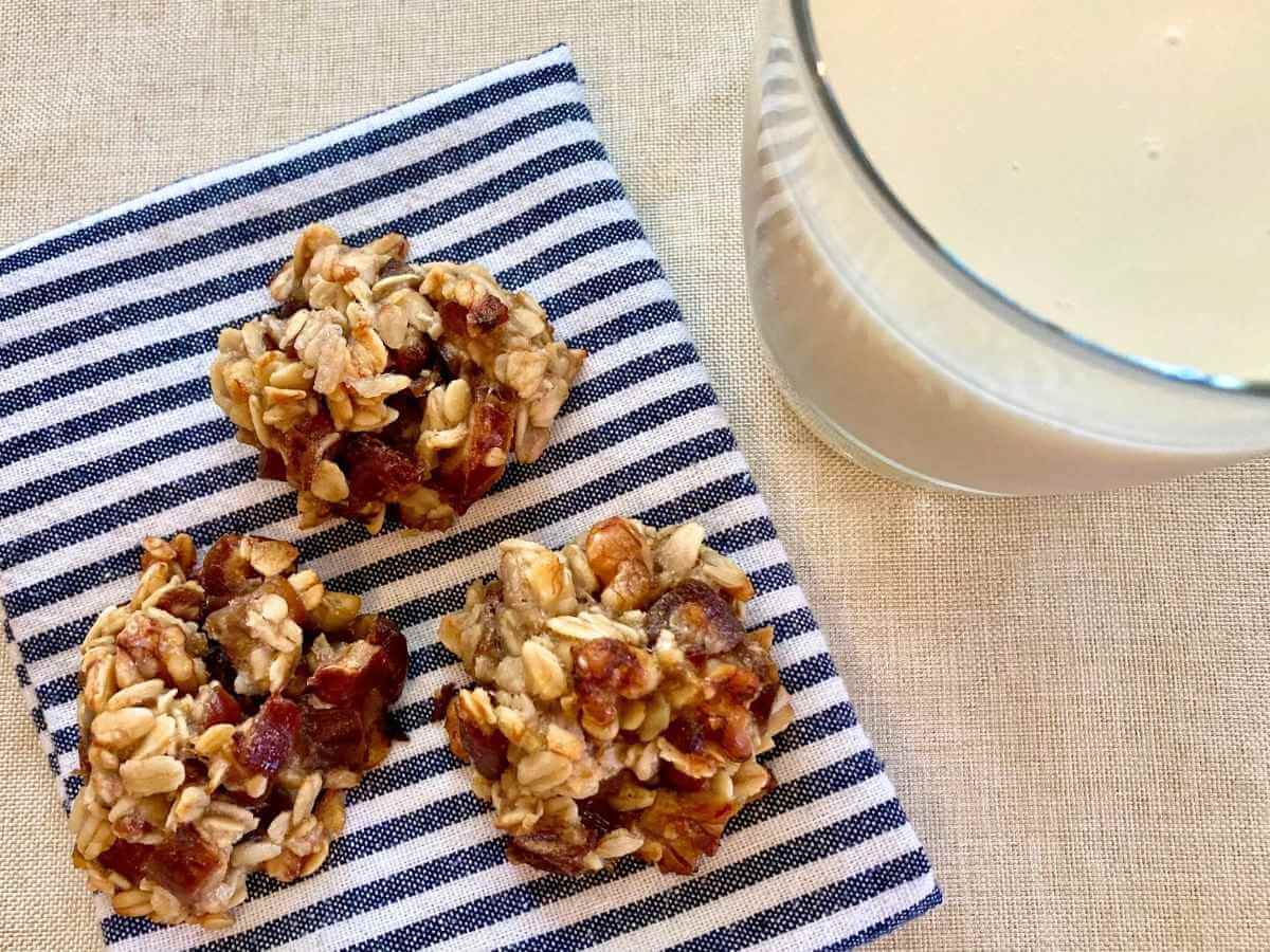 3 banana and date oatmeal cookies on a cloth napkin with a glass of milk.