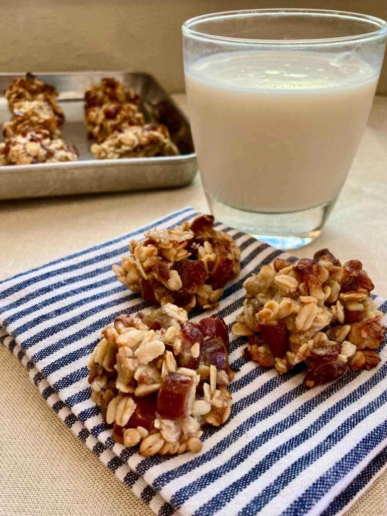 3 banana and date oatmeal cookies on a cloth napkin with a glass of milk and pan of cookies.