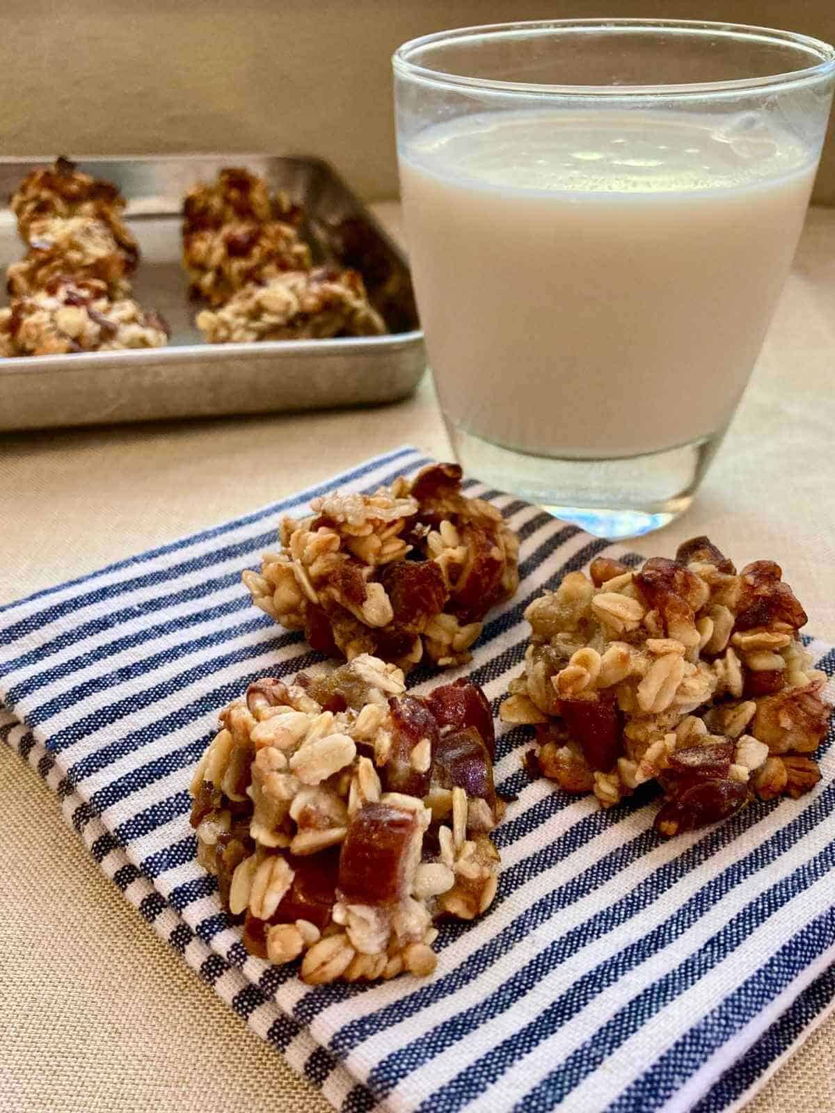 3 banana and date oatmeal cookies on a cloth napkin with a glass of milk and pan of cookies.