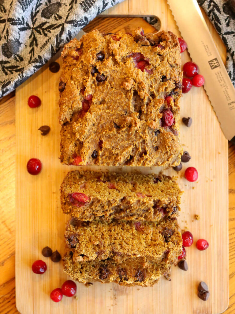 A partially sliced loaf of gluten-free pumpkin bread on a cutting board surrounded by cranberries and chocolate chips.