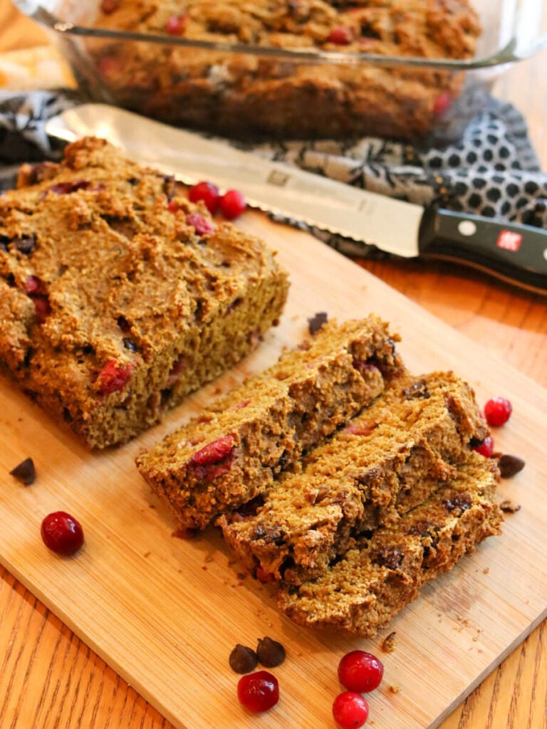 A partially sliced loaf of gluten-free pumpkin bread on a cutting board with a knife and another loaf of bread in the background.