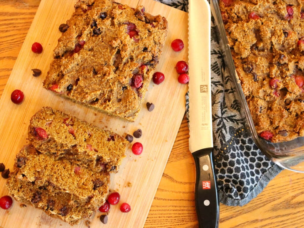 A partially sliced loaf of gluten-free pumpkin bread on a cutting board with a knife and a second loaf of bread in a bread pan.