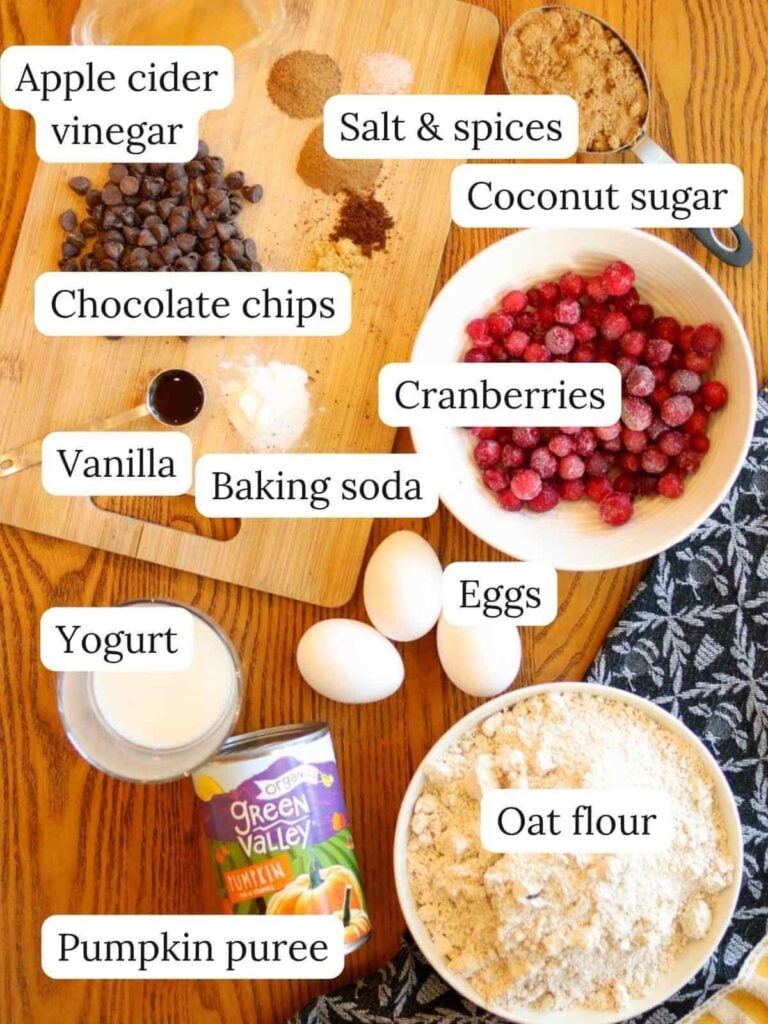 Labeled ingredients on a wooden table with a cutting board and dish towel including apple cider vinegar, salt, spices, coconut sugar, chocolate chips, vanilla, baking soda, cranberries, yogurt, eggs, pumpkin puree, and oat flour.