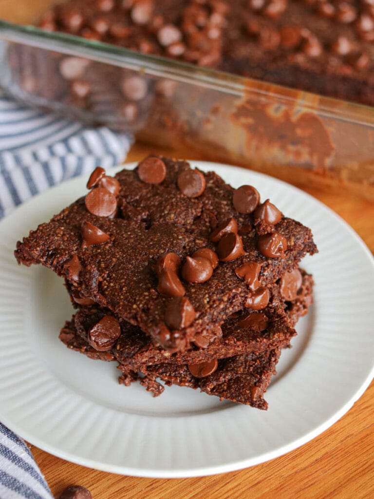 Stack of three vegan almond flour brownies with chocolate chips on a white plate with a pan of more brownies in the background.