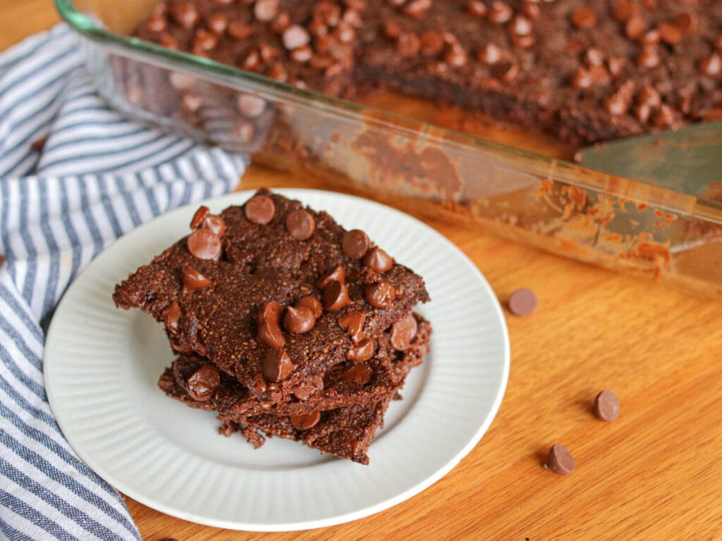 Stack of three vegan almond flour brownies with chocolate chips on a white plate on a wooden table with a pan of more brownies in the background.