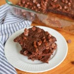 Stack of three vegan almond flour brownies with chocolate chips on a white plate on a wooden table with a cloth napkin with a pan of more brownies in the background.