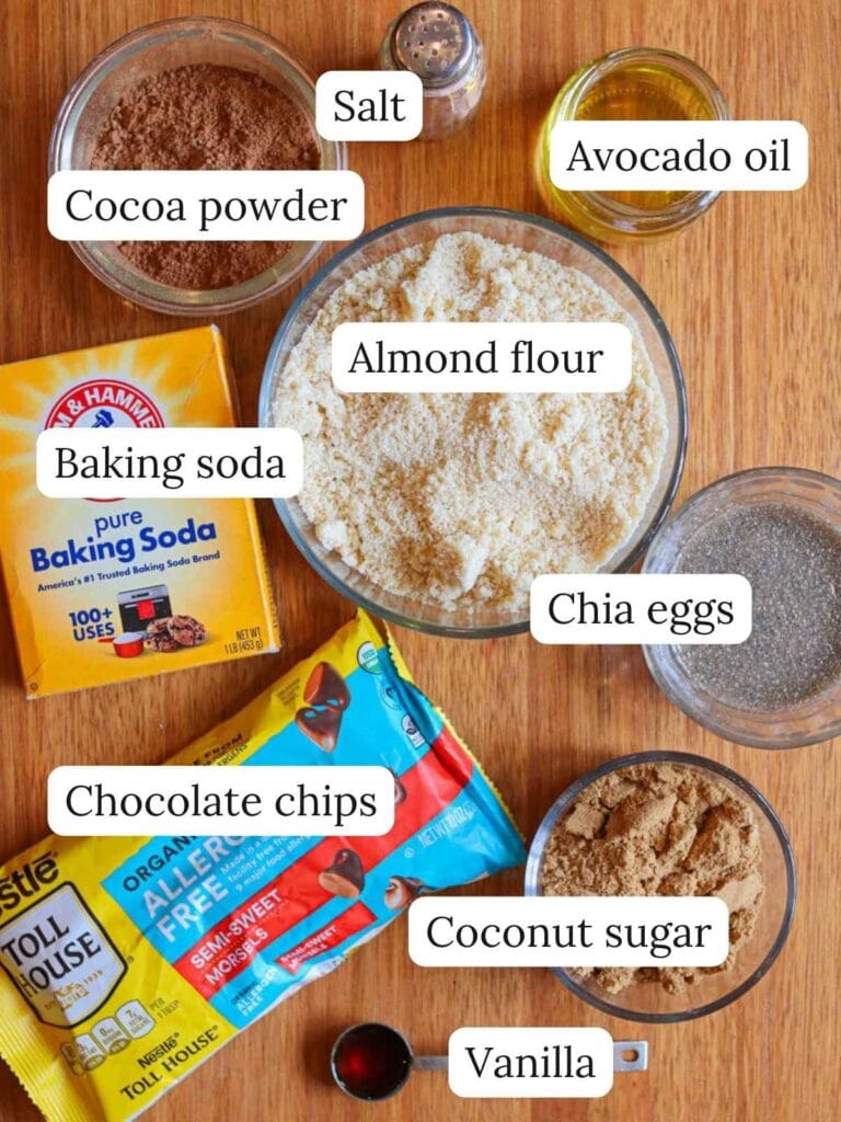 Labeled ingredients on a wooden surface including cocoa powder, salt, avocado oil, baking soda, almond flour, chia eggs, chocolate chips, coconut sugar, and vanilla.