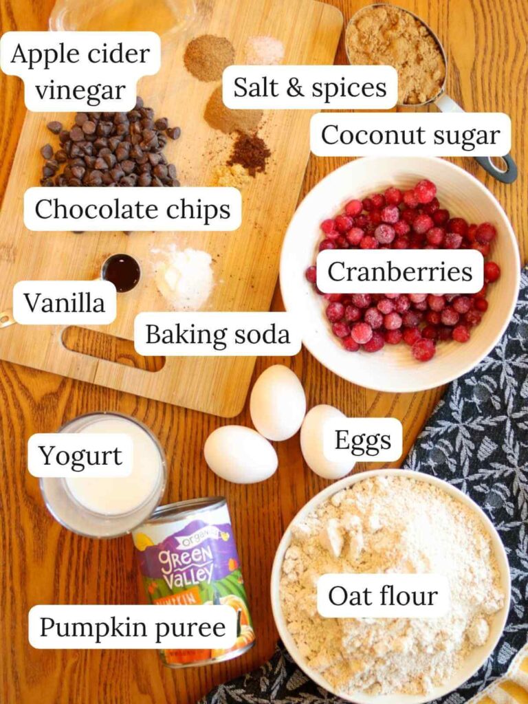 Labeled muffin ingredients on a wooden table with a cutting board and navy dish towel including apple cider vinegar, salt, spices, coconut sugar, chocolate chips, vanilla, baking soda, cranberries, yogurt, eggs, pumpkin puree, and oat flour.