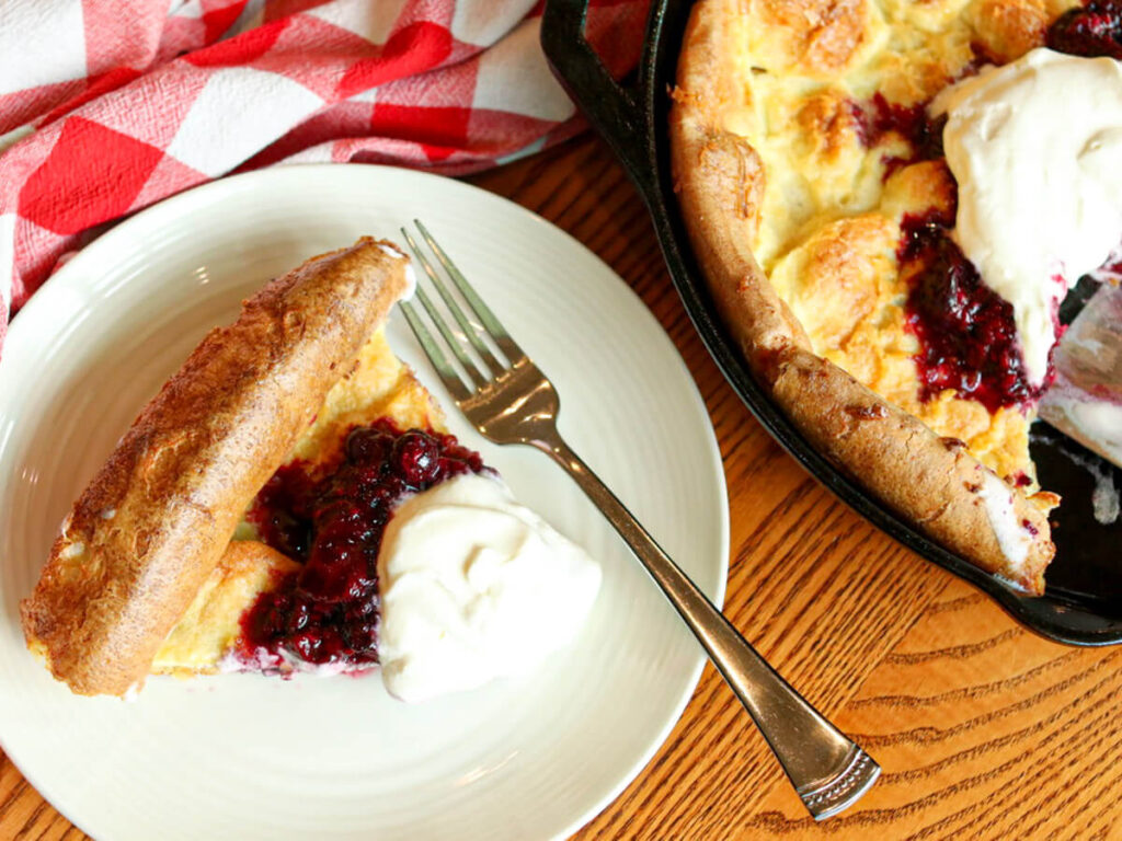 A slice of a Dutch baby on a plate topped with mixed berry compote and whipped cream beside a skillet with the rest of the Dutch baby.