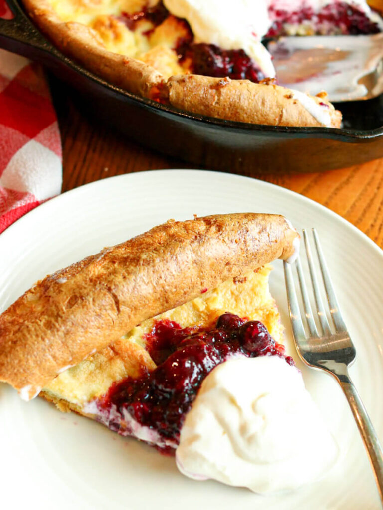 A slice of a Dutch baby on a plate with a fork topped with berries and whipped cream with more Dutch baby in the background.