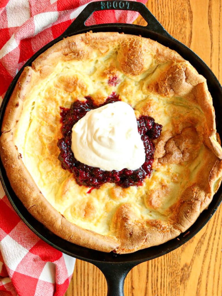 Oat flour Dutch baby in a skillet topped with berry compote and whipped cream on a wood table with a red and white cloth.