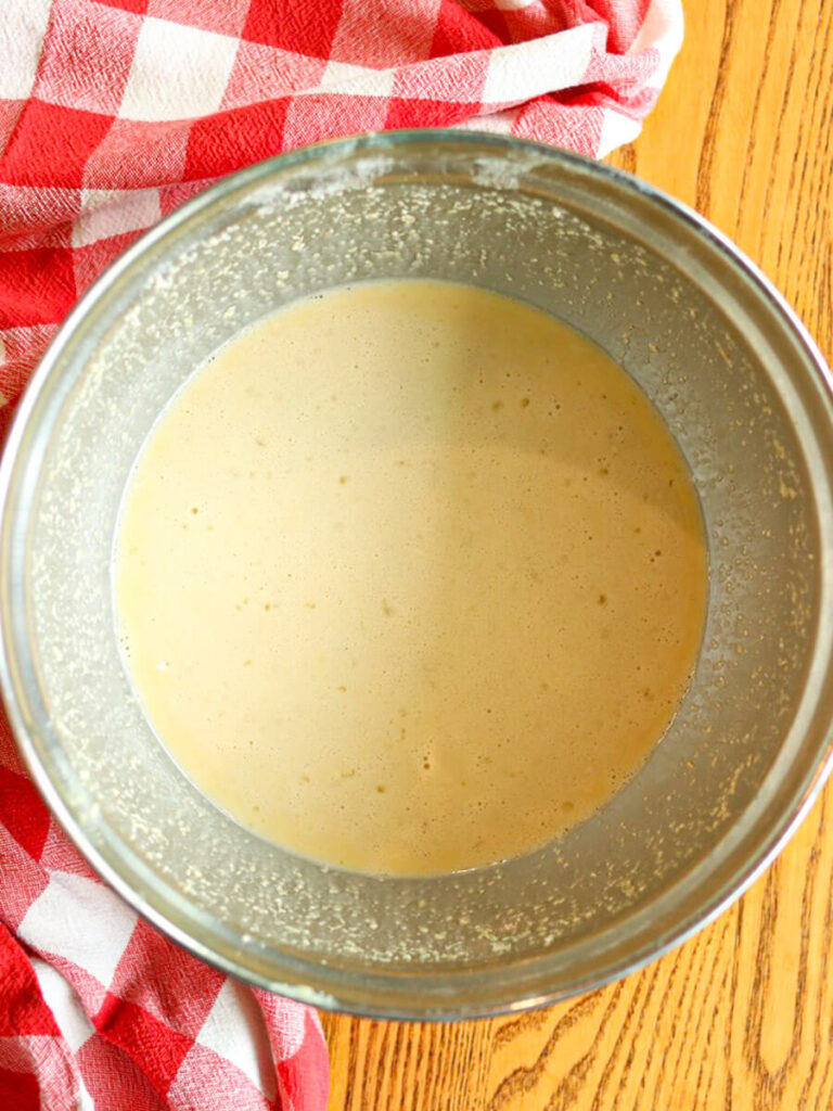 Dutch baby batter in a stainless steel mixing bowl on a wooden surface with a checkered cloth.
