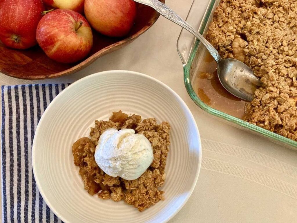 Apple crisp and ice cream in a bowl beside pan of apple crisp.