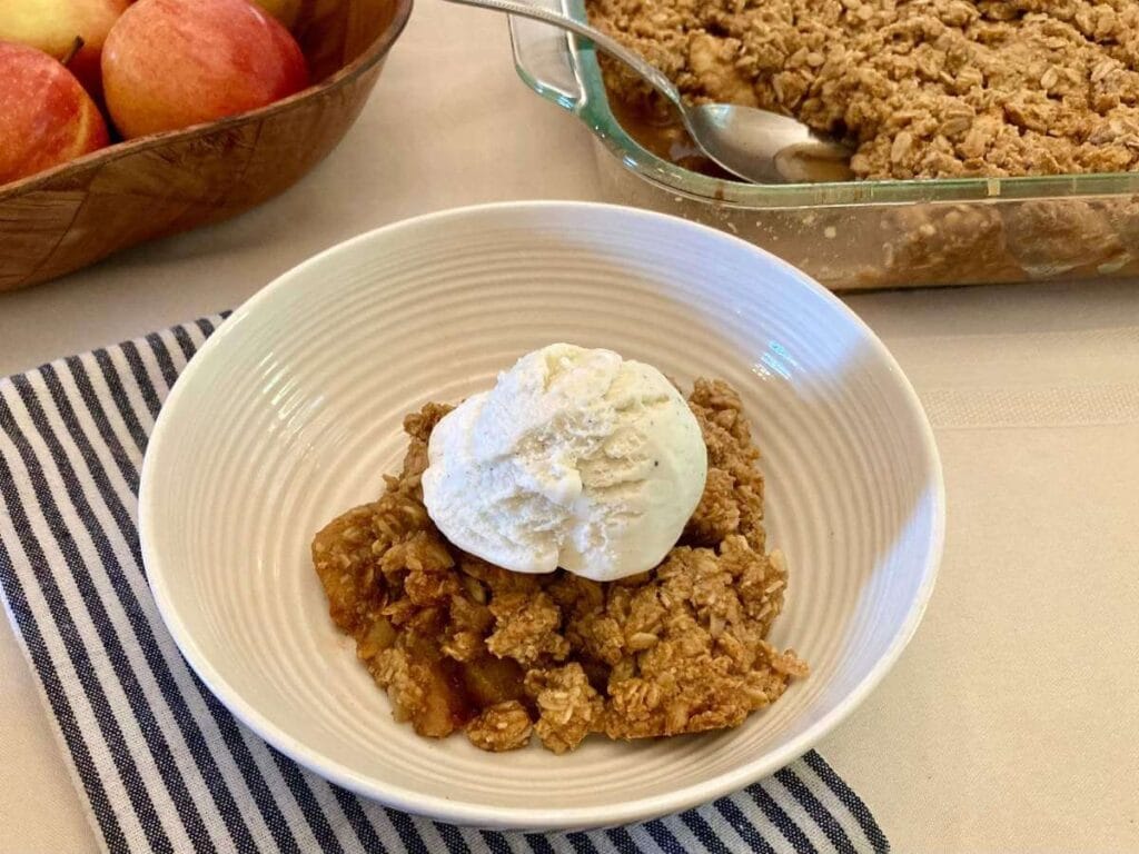 Apple crisp in a bowl with pan of apple crisp in the background.