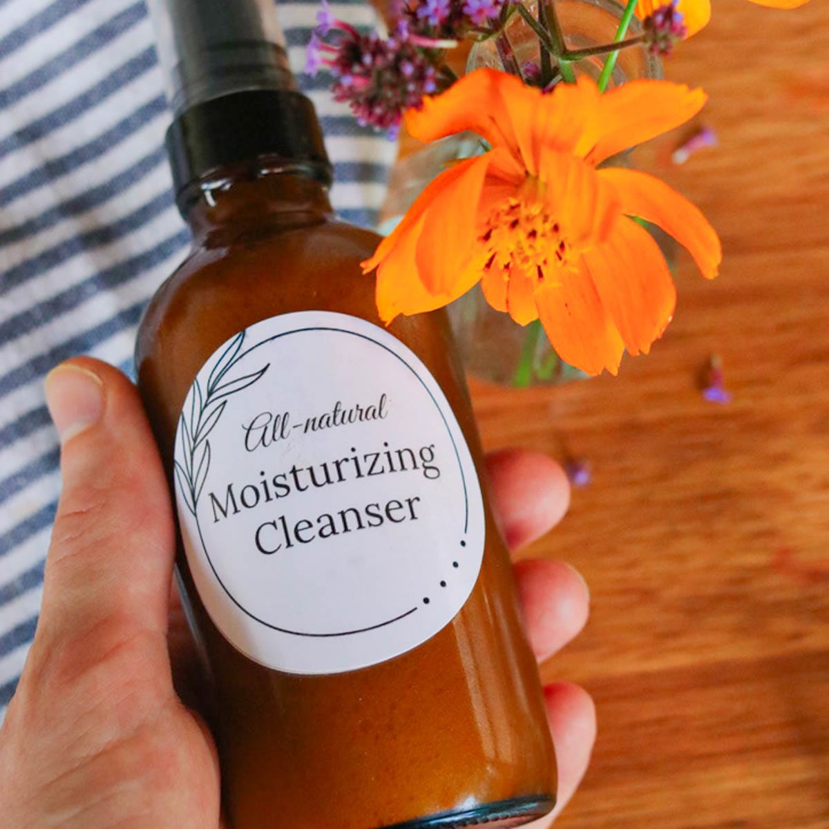 Hand holding amber glass spray bottle above a wooden table with a striped cloth and wildflowers.
