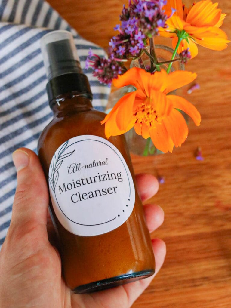 Hand holding amber glass spray bottle above a wooden table with a striped cloth and vase of wildflowers.