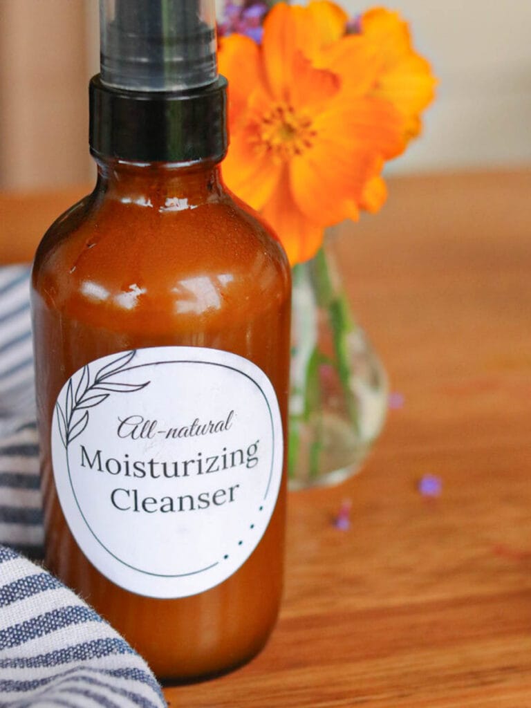 Amber glass spray bottle of face wash sitting on a wooden surface with a striped cloth a vase of wildflowers.
