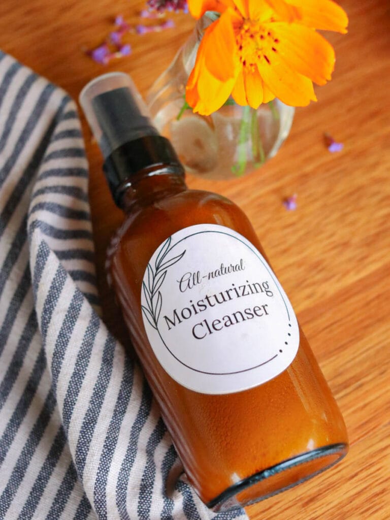 Amber glass spray bottle with DIY face wash on a wooden surface with a striped cloth a vase of wildflowers.