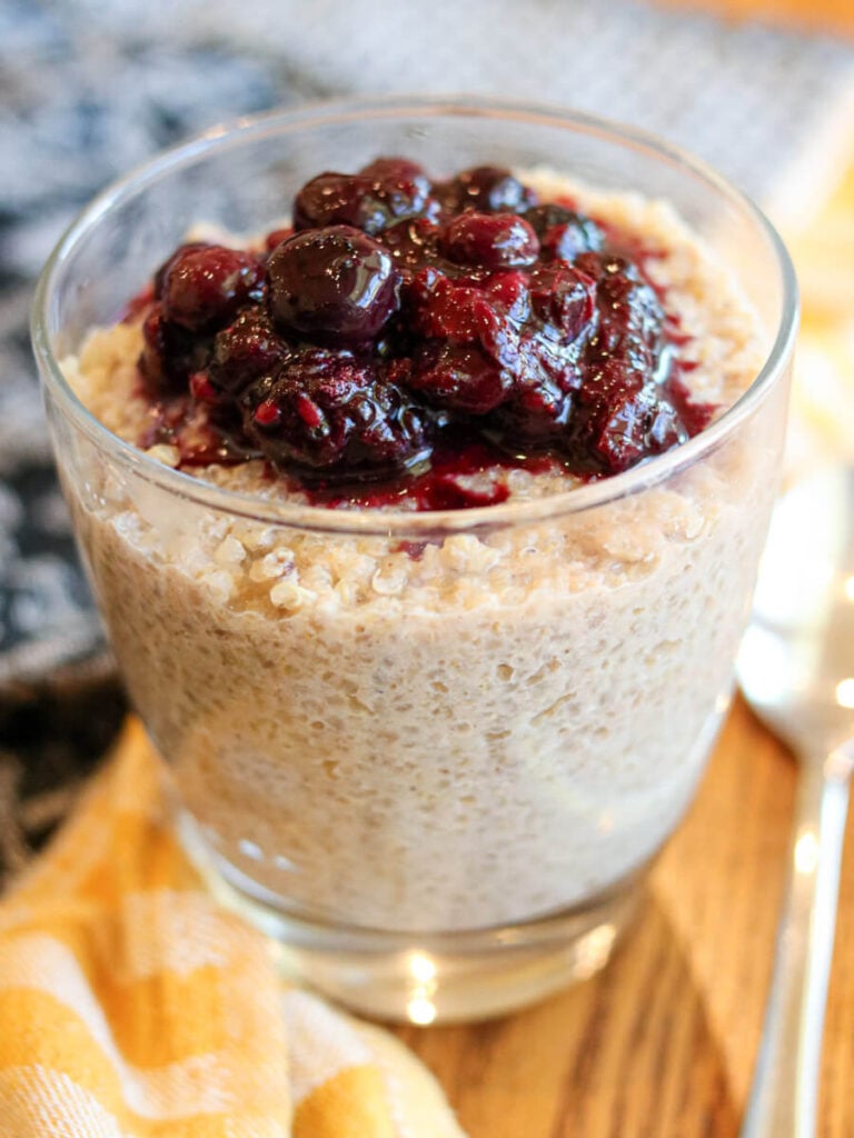 Glass dish full of quinoa pudding and berries on a wooden table with a dish cloth.