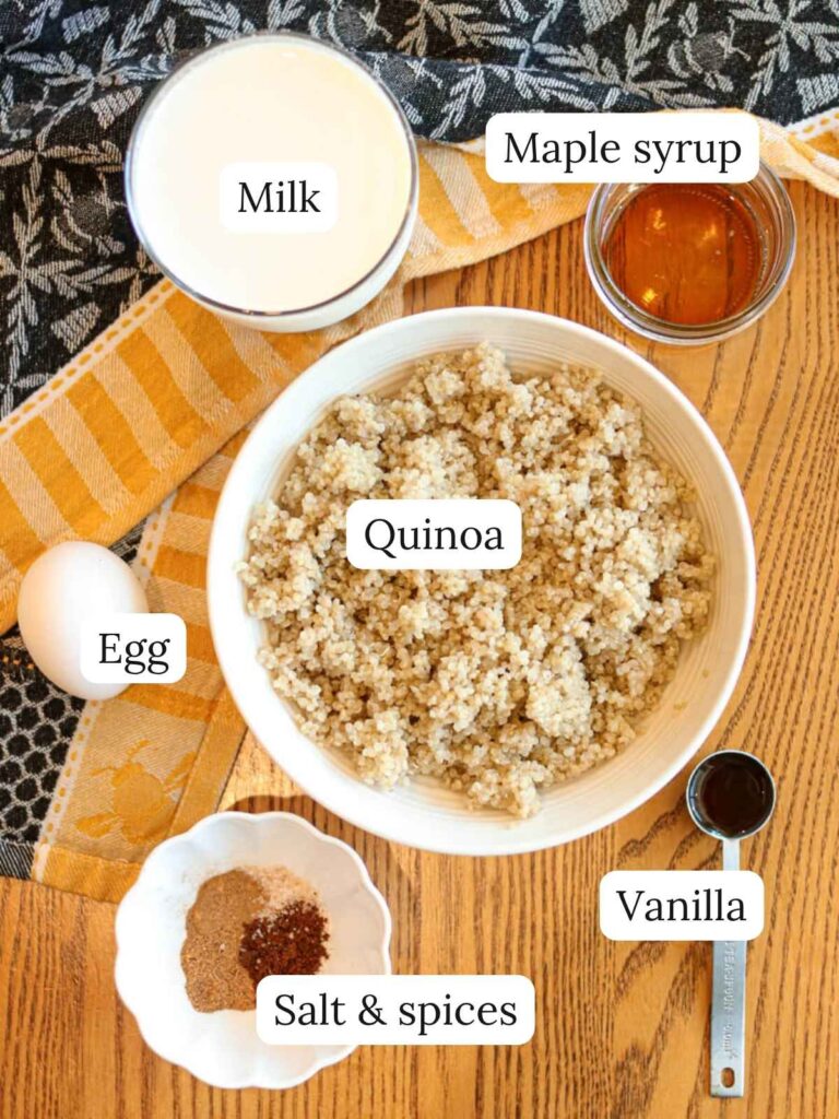 Labeled ingredients on a wood table with a yellow and navy dishcloth including milk, maple syrup, quinoa, egg, vanilla, salt, and spices.