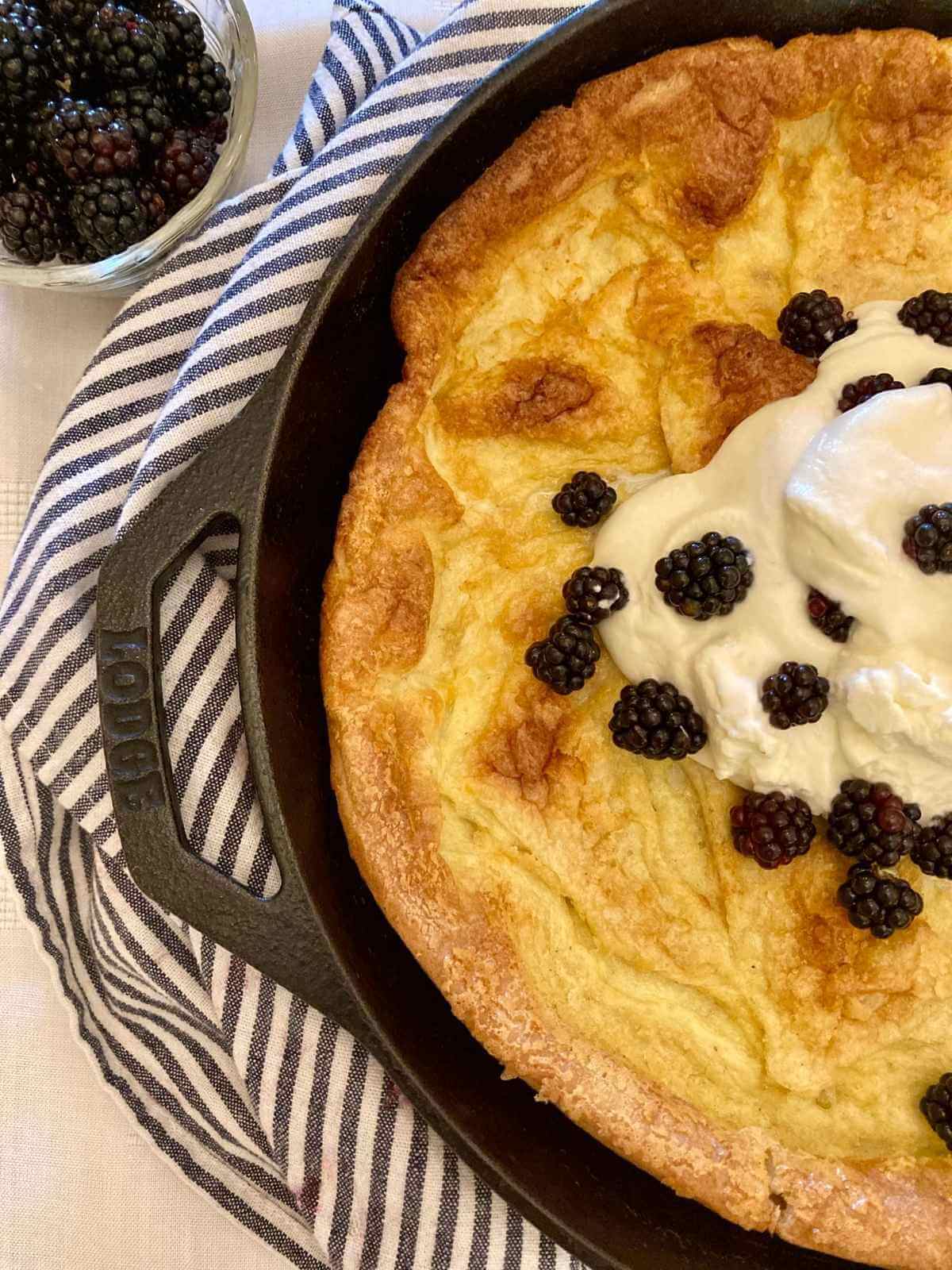 Closeup of half of a German pancake with whipped cream and blackberries in a skillet.
