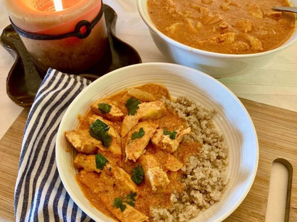 Indian chicken curry in a bowl with quinoa and in a serving bowl beside a candle.