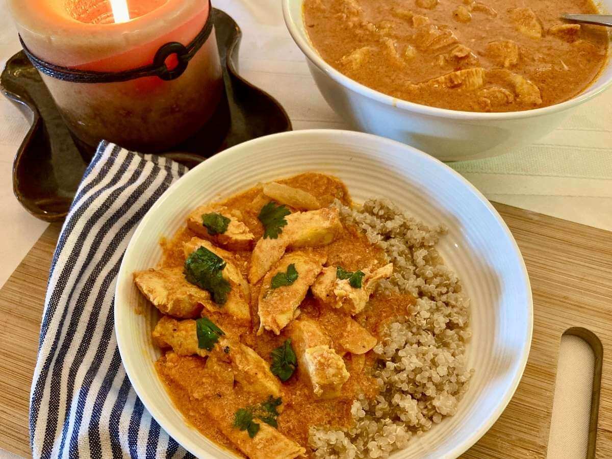 Indian chicken curry in a bowl with quinoa and in a serving bowl beside a candle.