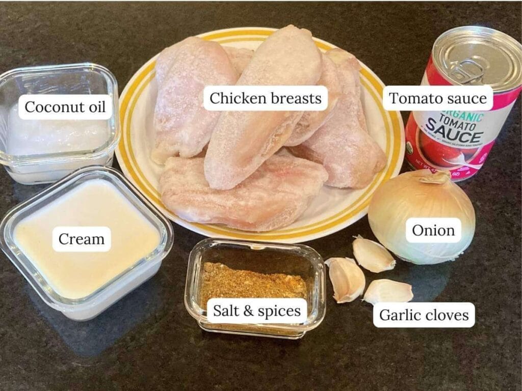 Ingredients for slow cooker chicken curry on a black surface including chicken breasts, tomato sauce, and spice.