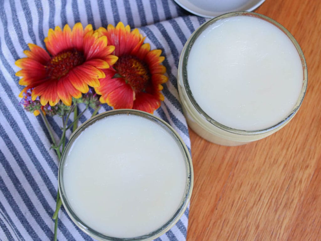 Two open glass jars of tallow balm on a wooden surface with a blue striped cloth and wildflowers.