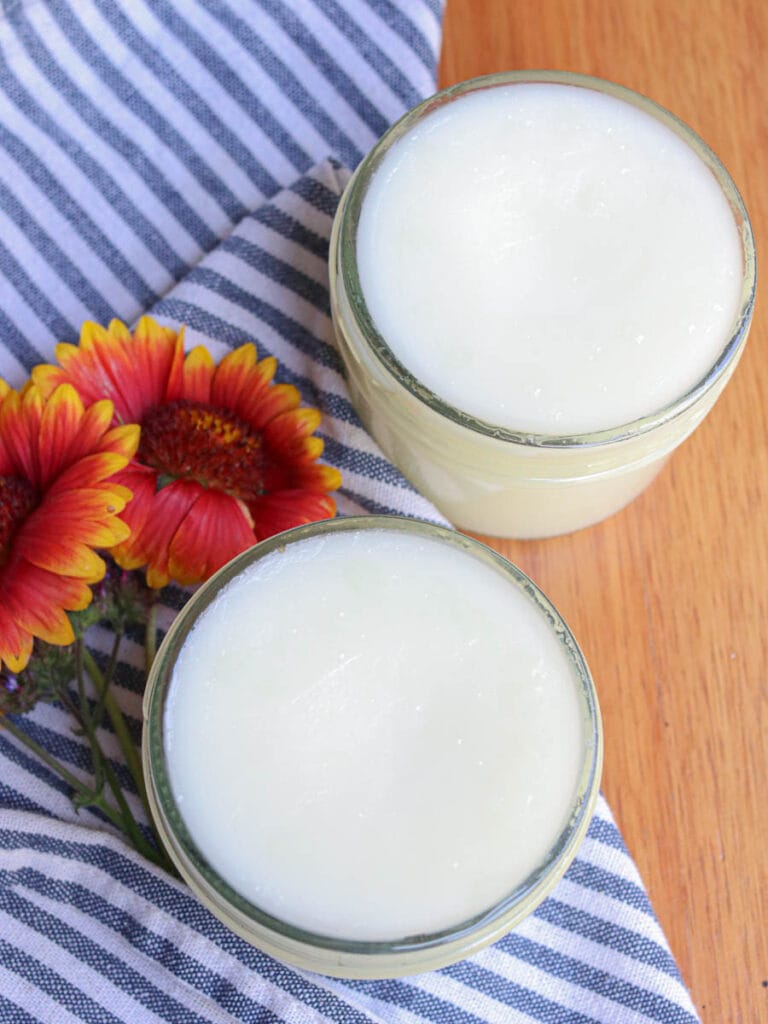 Two open glass jars of tallow balm on a wooden surface with a striped cloth and fresh flowers.