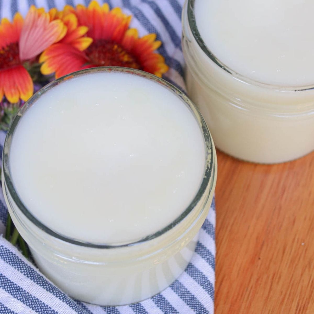 Two open glass jars of tallow balm on a wooden surface with a striped cloth and flowers.