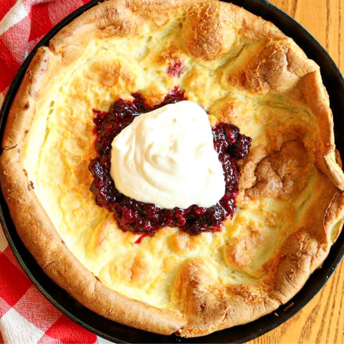 Closeup view of a Dutch baby in a skillet topped with berry sauce and homemade whipped cream.
