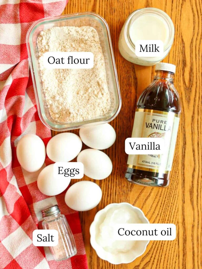 Labeled ingredients on a wooden table with a checkered cloth including oat flour, milk, vanilla, eggs, salt, and coconut oil.