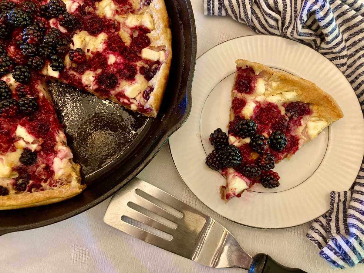 Slice of blackberry cheesecake German pancakes on a plate with a skillet with the rest of the pancake.