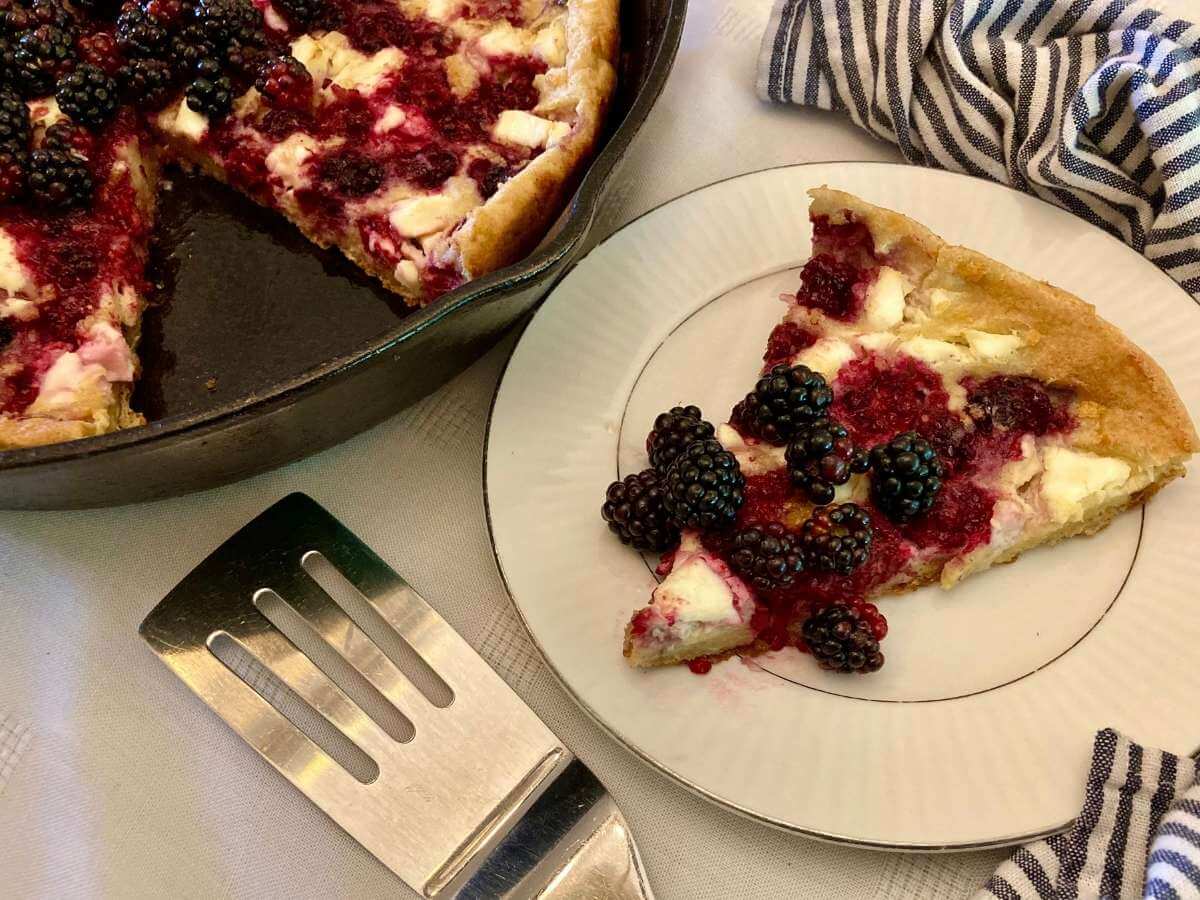 Slice of blackberry cheesecake German pancakes on a plate with a skillet with the rest of the pancake in the background.