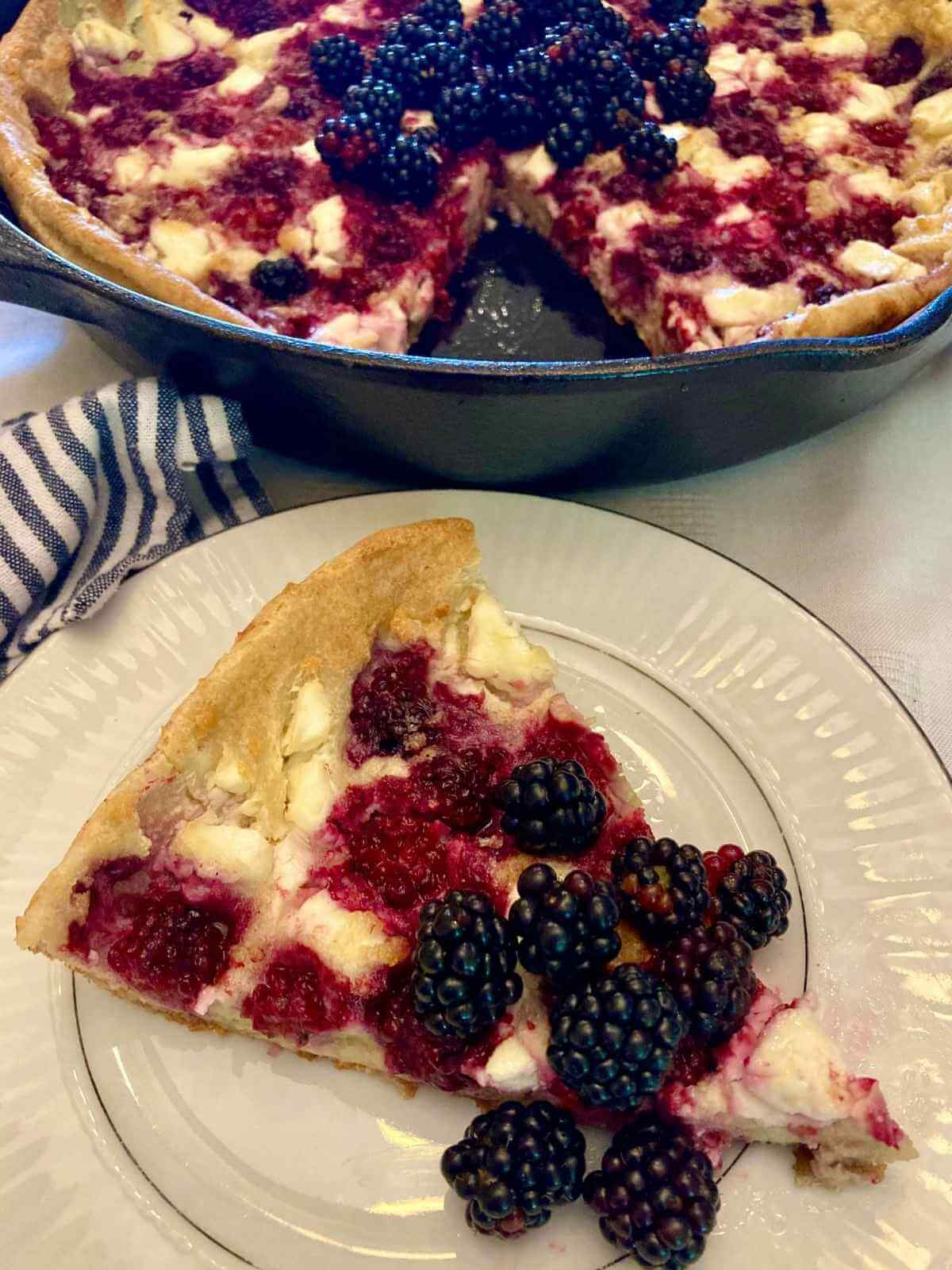 Closeup of slice of blackberry cheesecake German pancakes on a plate with a skillet with the rest of the pancake.