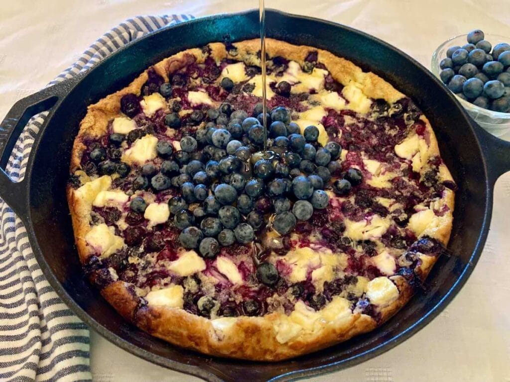 Skillet with blueberry cheesecake German pancake with blueberries on top and drizzled with syrup.