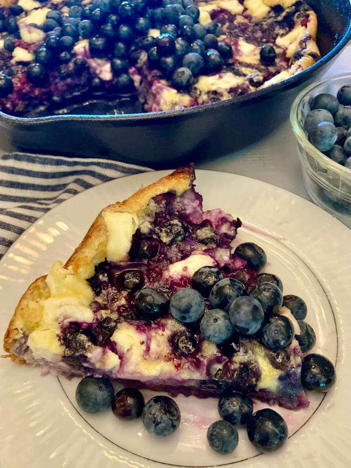Slice of blueberry cheesecake German pancake on a plate with the rest of the pancake in the background.