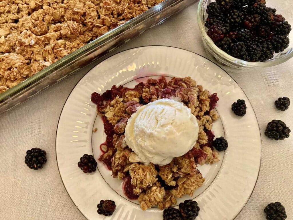 Gluten-free blackberry crisp and ice cream on a plate beside pan of blackberry crisp.