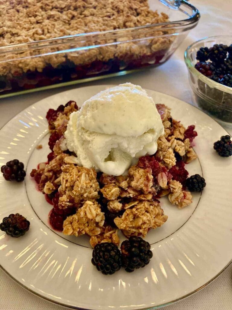 Gluten-free blackberry crisp with ice cream on a plate with pan of blackberry crisp in the background.