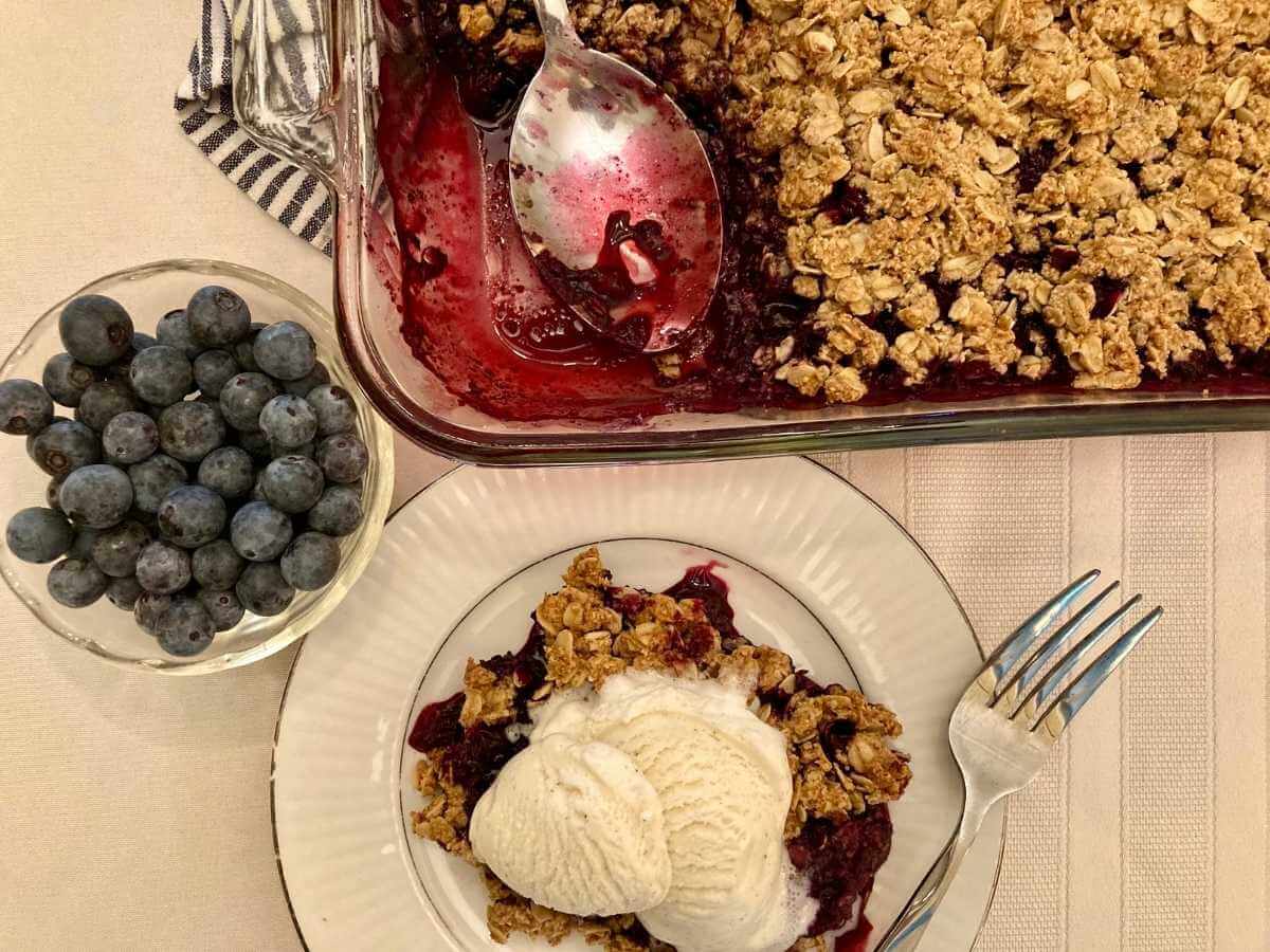 Gluten-free blueberry crisp with ice cream on a plate with pan of blueberry crisp.