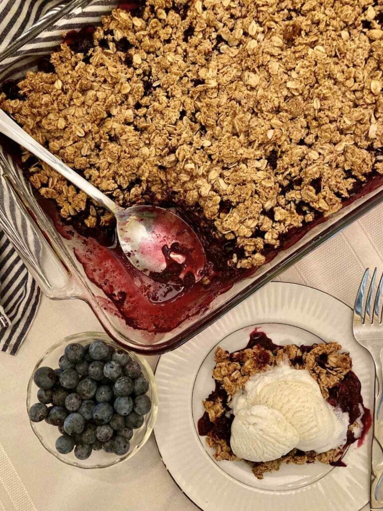 Gluten-free blueberry crisp in a glass baking dish and vanilla ice cream on a plate beside pan of blueberry crisp.
