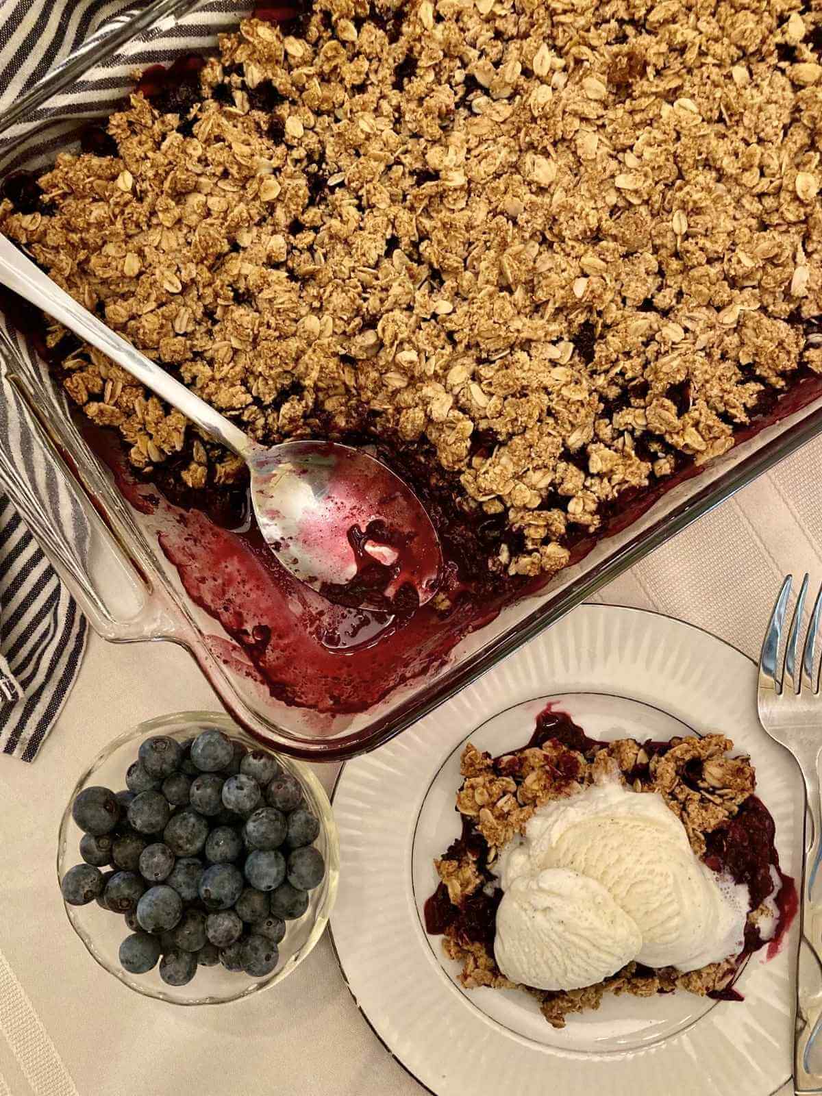Gluten-free blueberry crisp and vanilla ice cream on a plate beside pan of blueberry crisp.