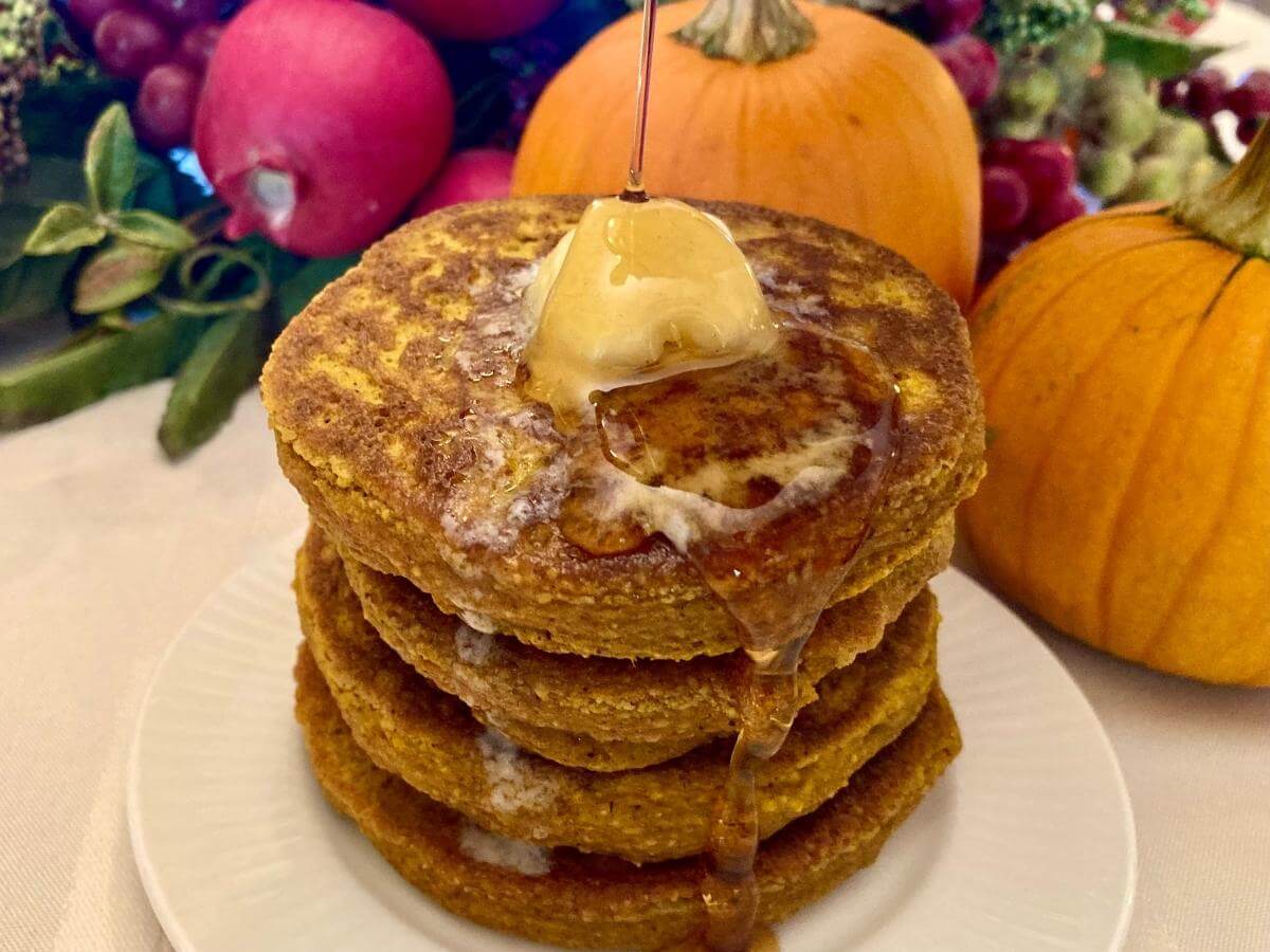 Pouring syrup over a stack of gluten-free pumpkin pancakes on a plate.