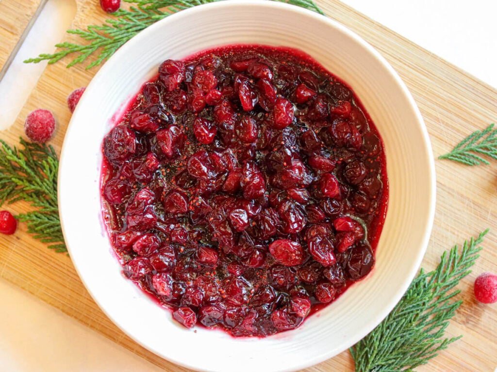 Healthy cranberry sauce in a white bowl on a cutting board surrounded by evergreen leaves and cranberries.