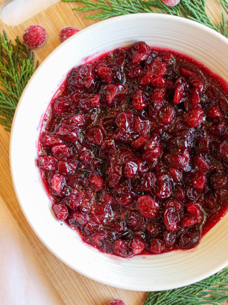 Closeup view of cranberry sauce in a serving bowl on a wooden surface with evergreen leaves and fresh cranberries.