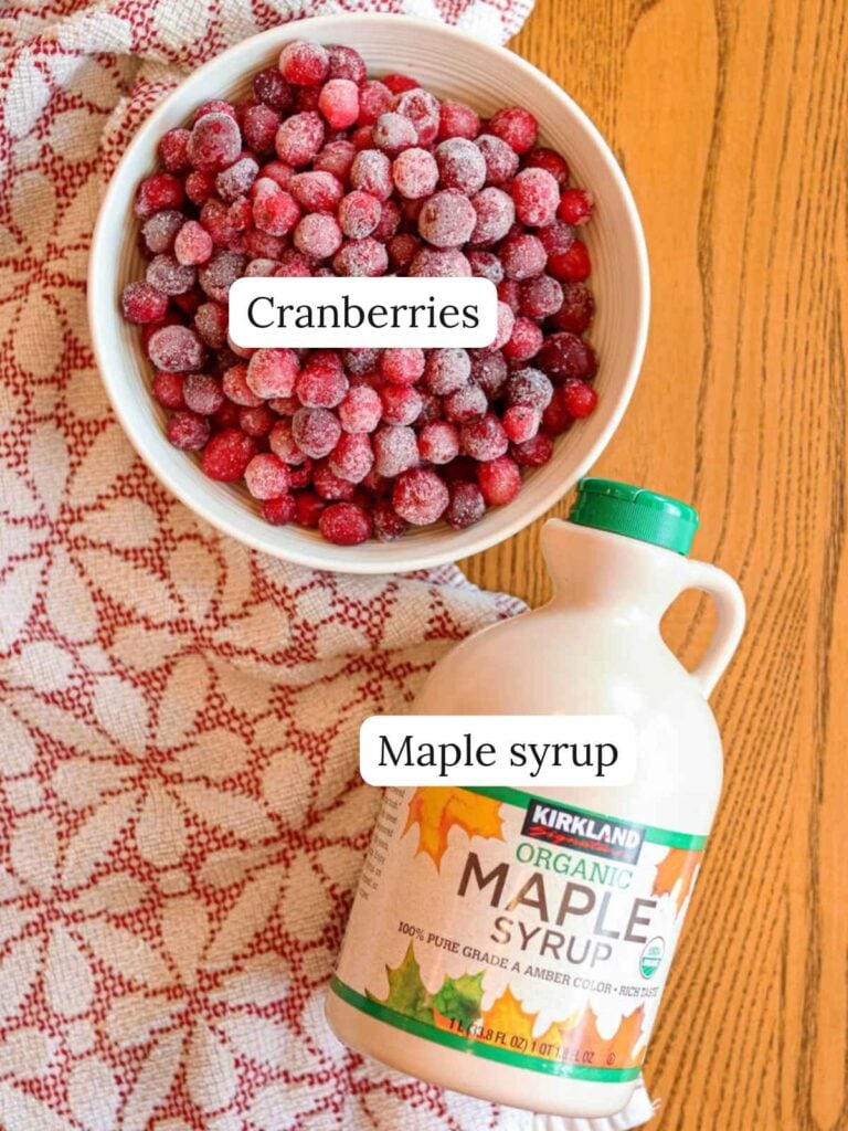 Labeled ingredients on a wood table with a kitchen towel including cranberries and maple syrup.