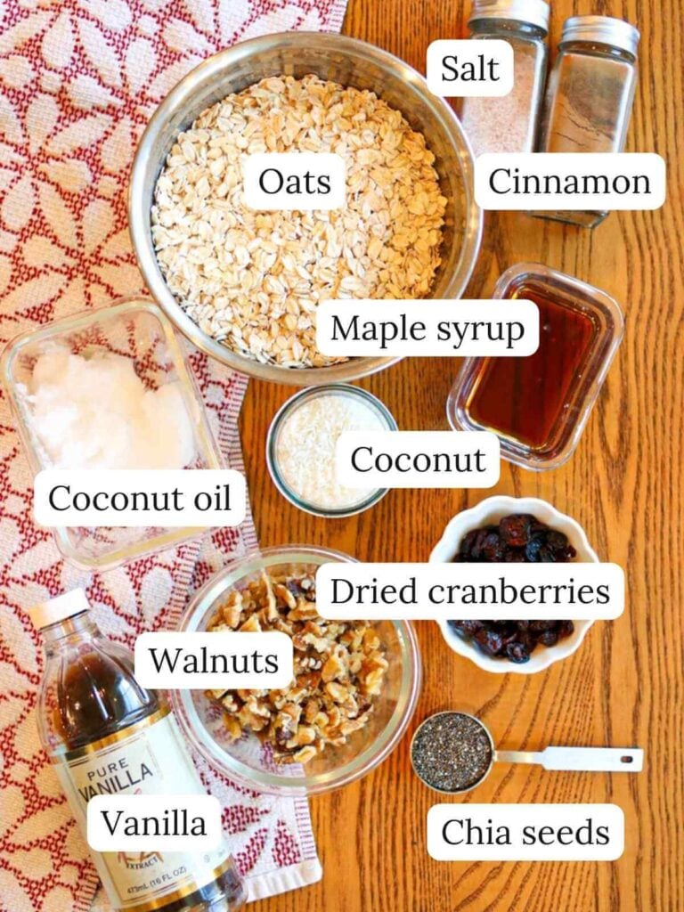 Labeled ingredients on a wooden table including oats, salt, cinnamon, coconut oil, coconut, maple syrup, vanilla, walnuts, dried cranberries, and chia seeds.