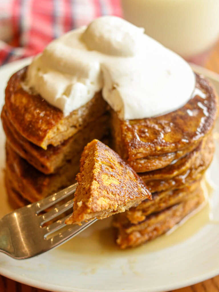 A stack of oatmeal pumpkin pancakes topped with whipped cream with a cut in them and a bite on a fork.