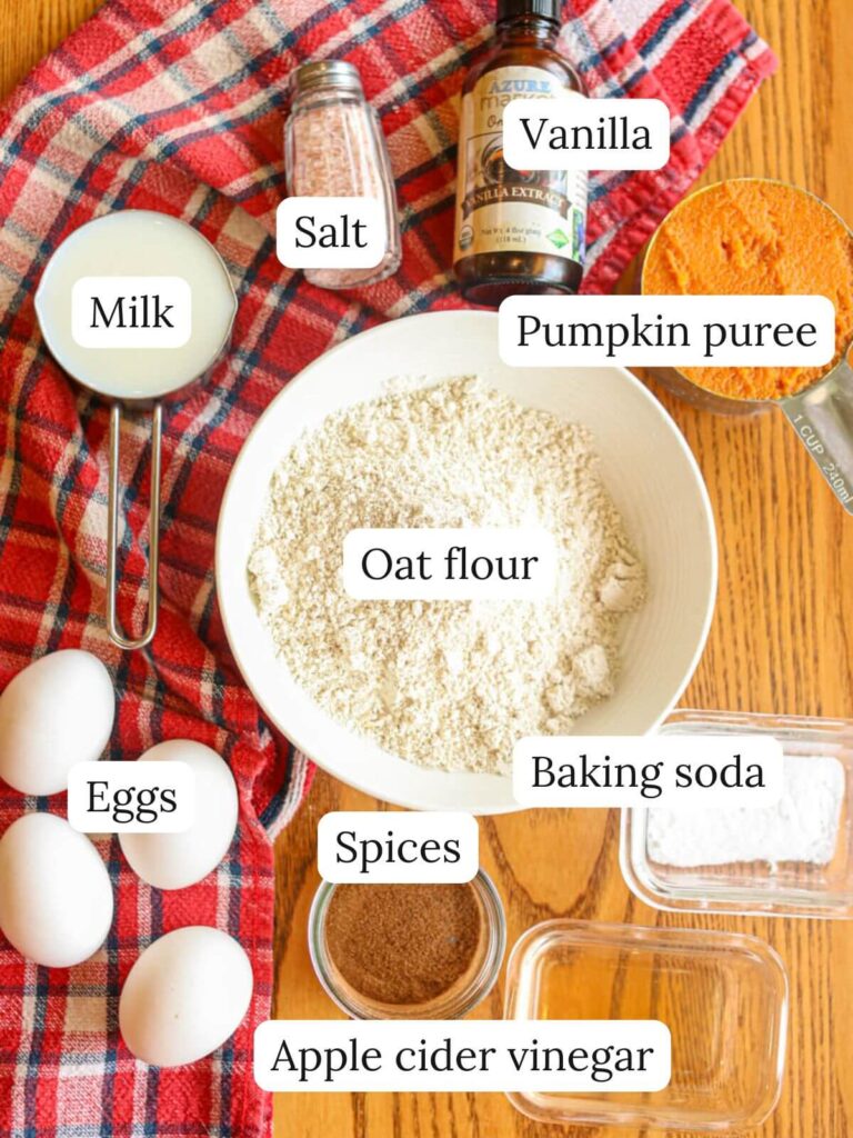 Labeled ingredients on a table with a kitchen towel including oat flour, milk, eggs, pumpkin puree, spices, baking soda, apple cider vinegar, vanilla, and salt.
